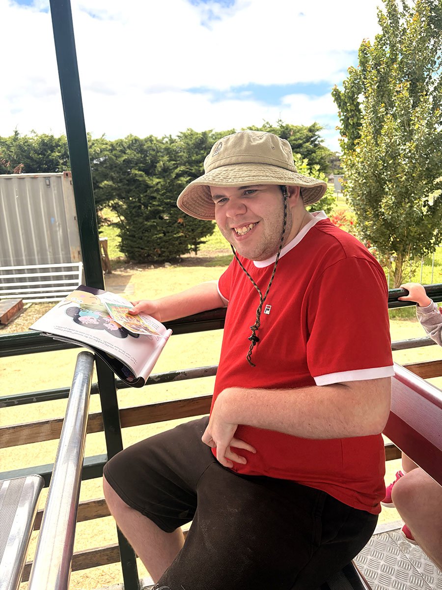 A man wearing a beige sun hat and a red T-shirt, smiling while sitting outdoors on a sunny day, reading a magazine.