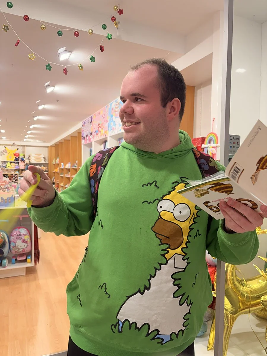 A man smiling at a store, wearing a bright green hoodie with a cartoon character from The Simpsons printed on it. He holds a brochure in one hand and a yellow object in the other. The store has Christmas decorations hanging from the ceiling.
