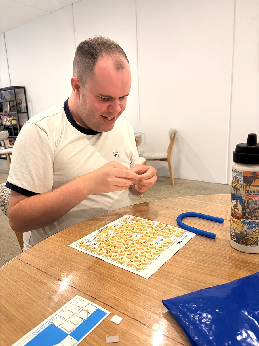 A man sitting at a wooden table playing bingo, holding an orange bingo marker and looking at the bingo card. There is a blue bingo marker, a water bottle with colorful travel stickers, and a blue plastic bag on the table inside a room with white wall