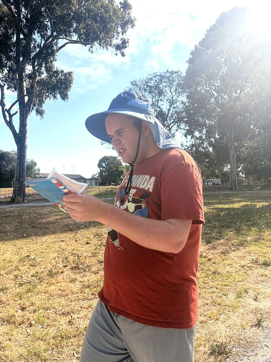 A young man outdoors in bright sunlight, wearing a wide-brimmed hat, red T-shirt with cartoon graphic, and gray shorts, reading a magazine in a park with trees and grassy area.