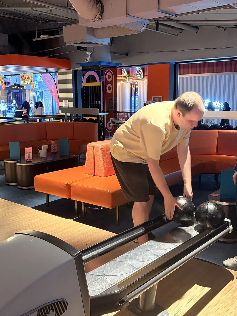 A man arranging bowling balls on a ball return rack at a bowling alley. The alley has orange and blue seating with a contemporary decor.