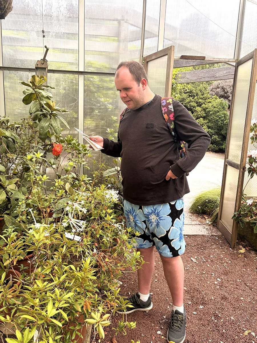 A man wearing a black sweatshirt, floral shorts, and sneakers is in a greenhouse, looking at and reading a small booklet or plant label, with his left hand in his pocket.