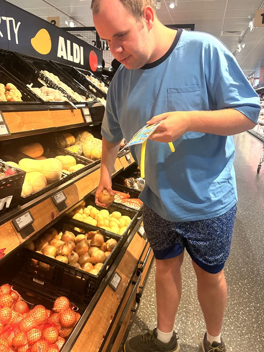 A man shopping for onions at an ALDI grocery store, holding a yellow plastic bag and selecting onions from a display shelf that has various types of onions and other produce.