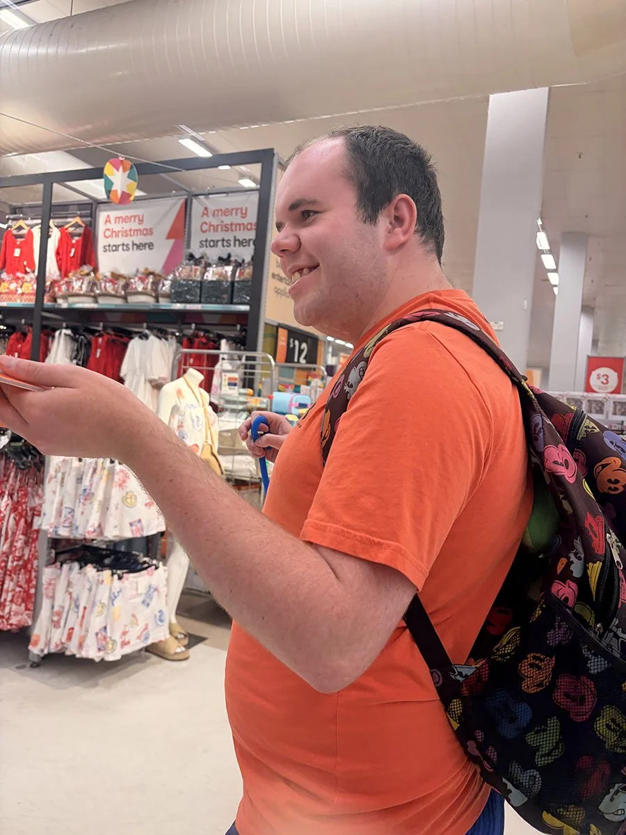 A smiling man with short dark hair wearing an orange shirt and a colorful backpack, shopping in a retail store with Christmas clothing and signs in the background.