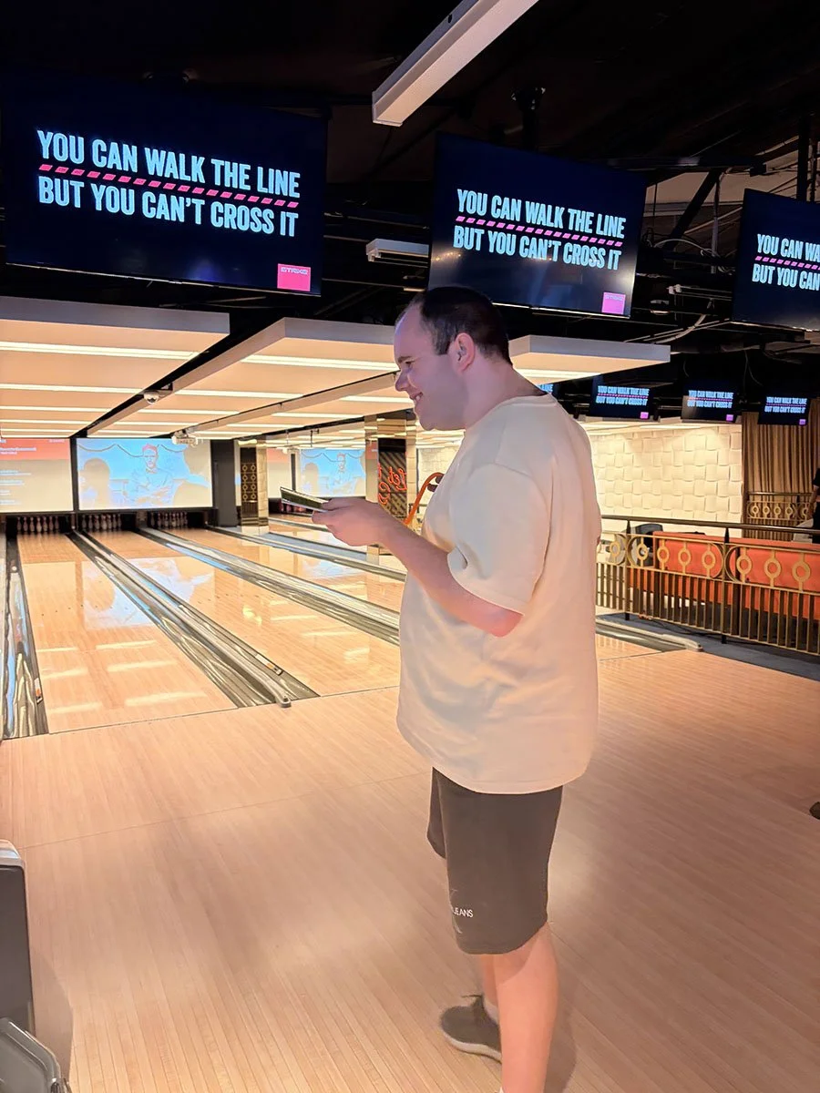 A young man standing in a bowling alley, looking at his phone, with multiple screens overhead displaying the message, "You can walk the line but you can't cross it."