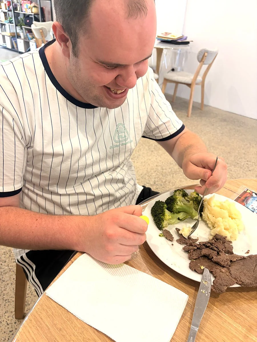 A man sitting at a table, smiling while eating a meal of broccoli, mashed potatoes, and slices of cooked beef.