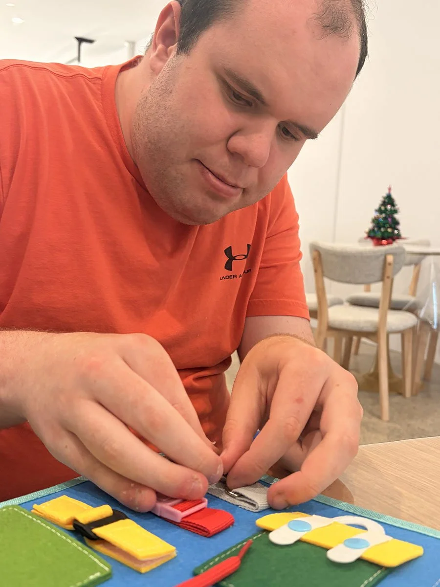 A man with short dark hair wearing a red Under Armour shirt, sitting at a table, concentrating on a tabletop activity involving small fabric pieces, possibly a craft or game. In the background, a small decorated Christmas tree is visible on a table, 