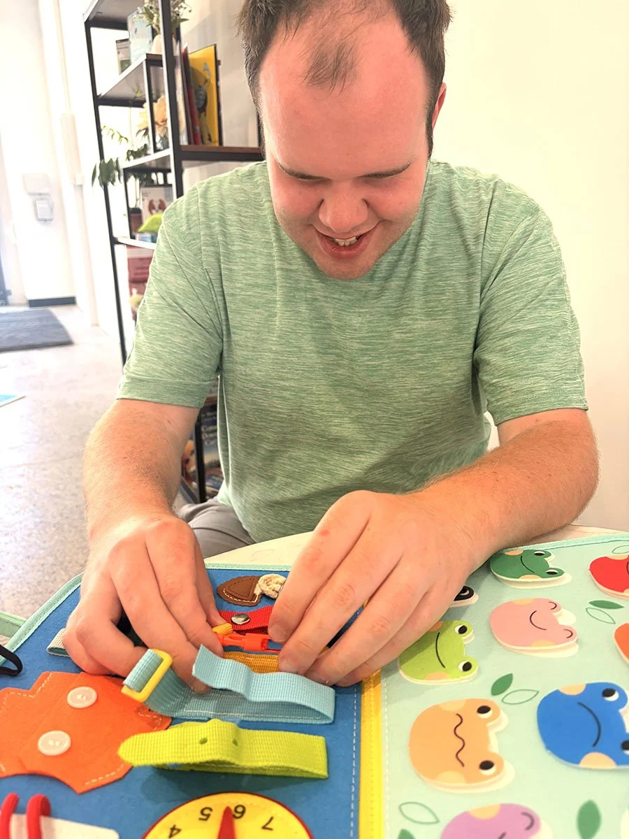 A man in a green shirt sitting at a table, playing with a colorful fabric activity book with animal faces and buttons.