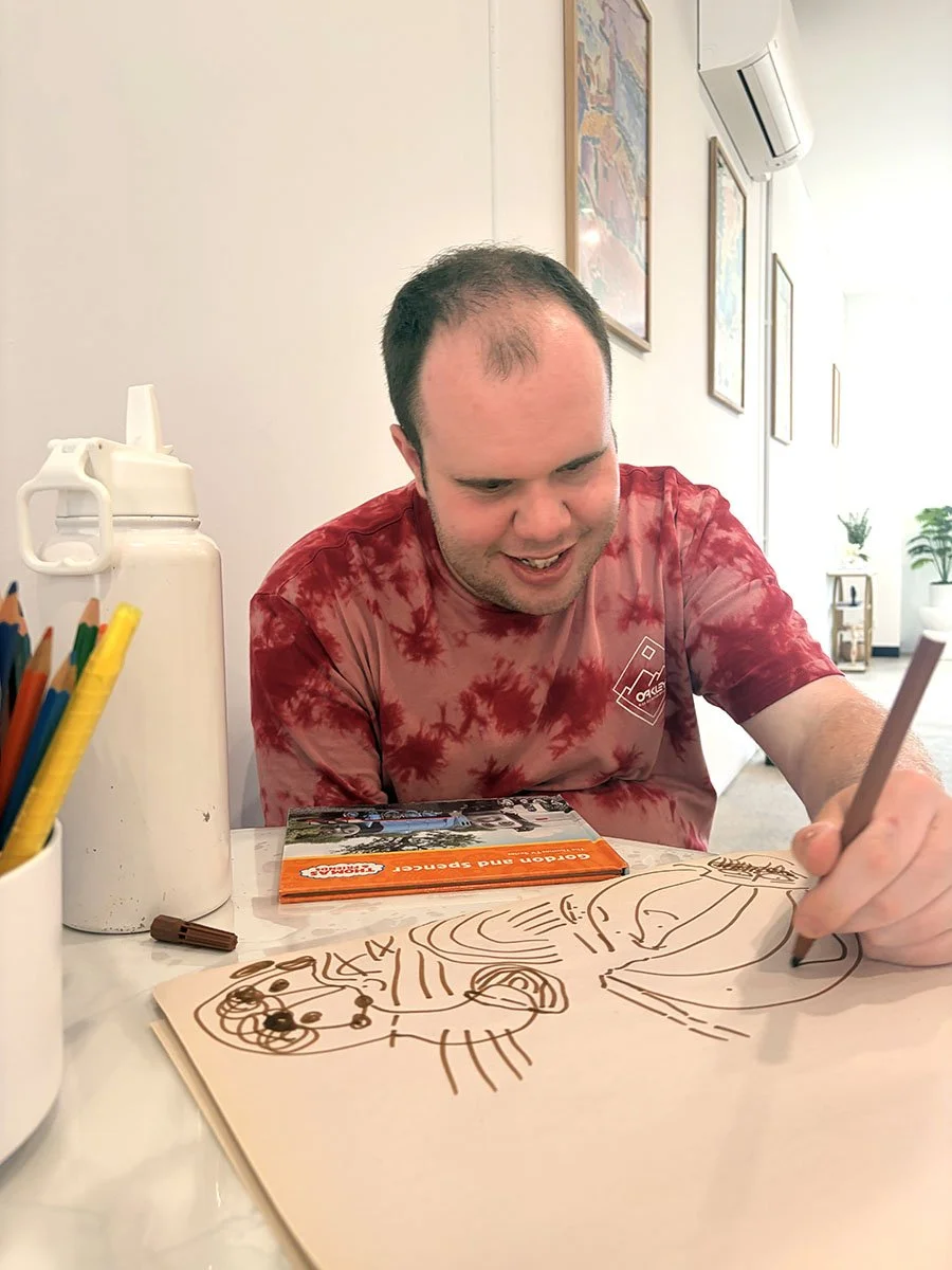 A man in a red tie-dye shirt drawing with a brown marker on a large sheet of paper, which has various cartoon-style sketches.