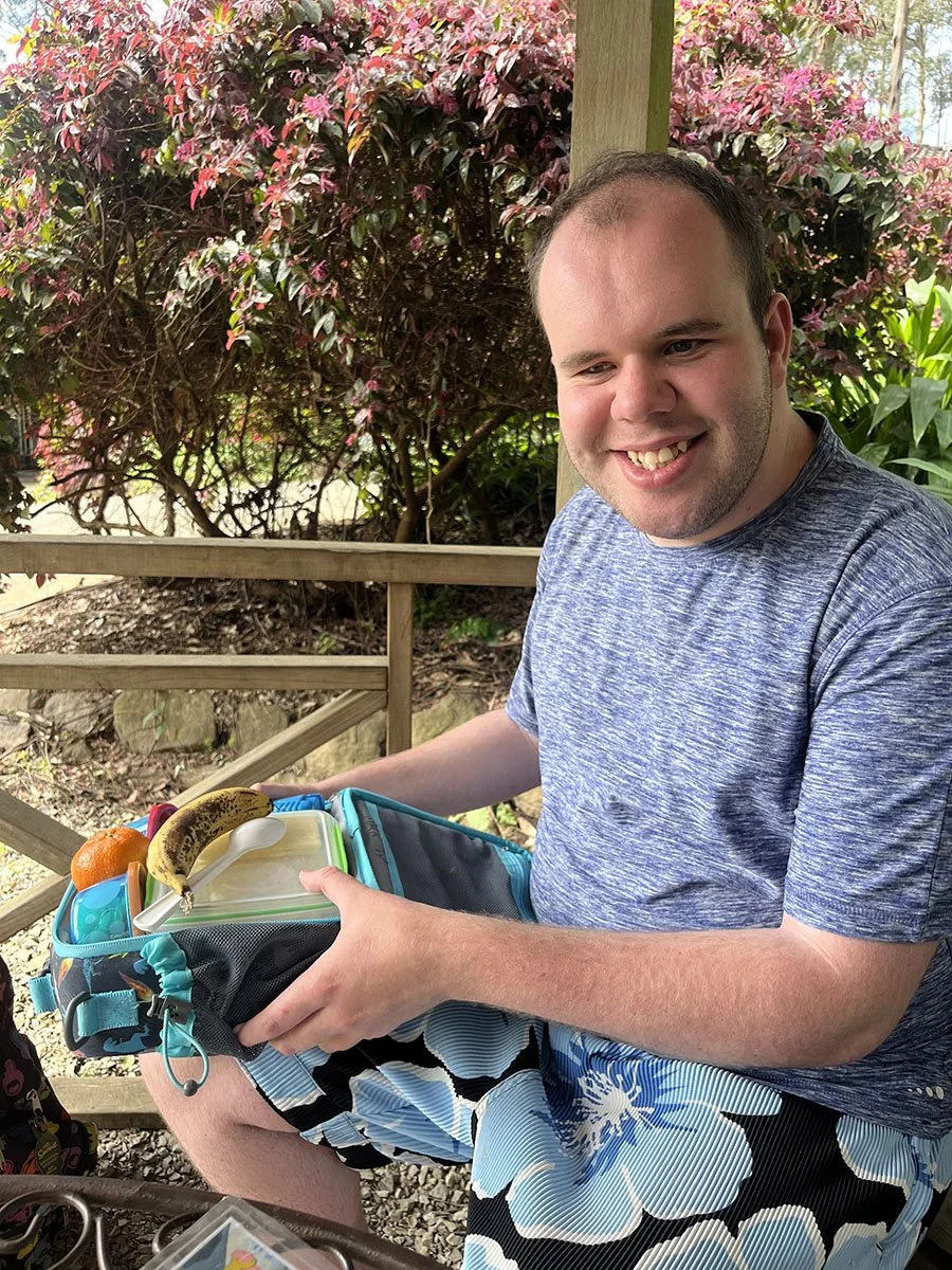 A man smiling outdoors, sitting on a wooden bench, holding a lunch box with a banana, tangerine, and other items, with pink flowering bushes in the background.