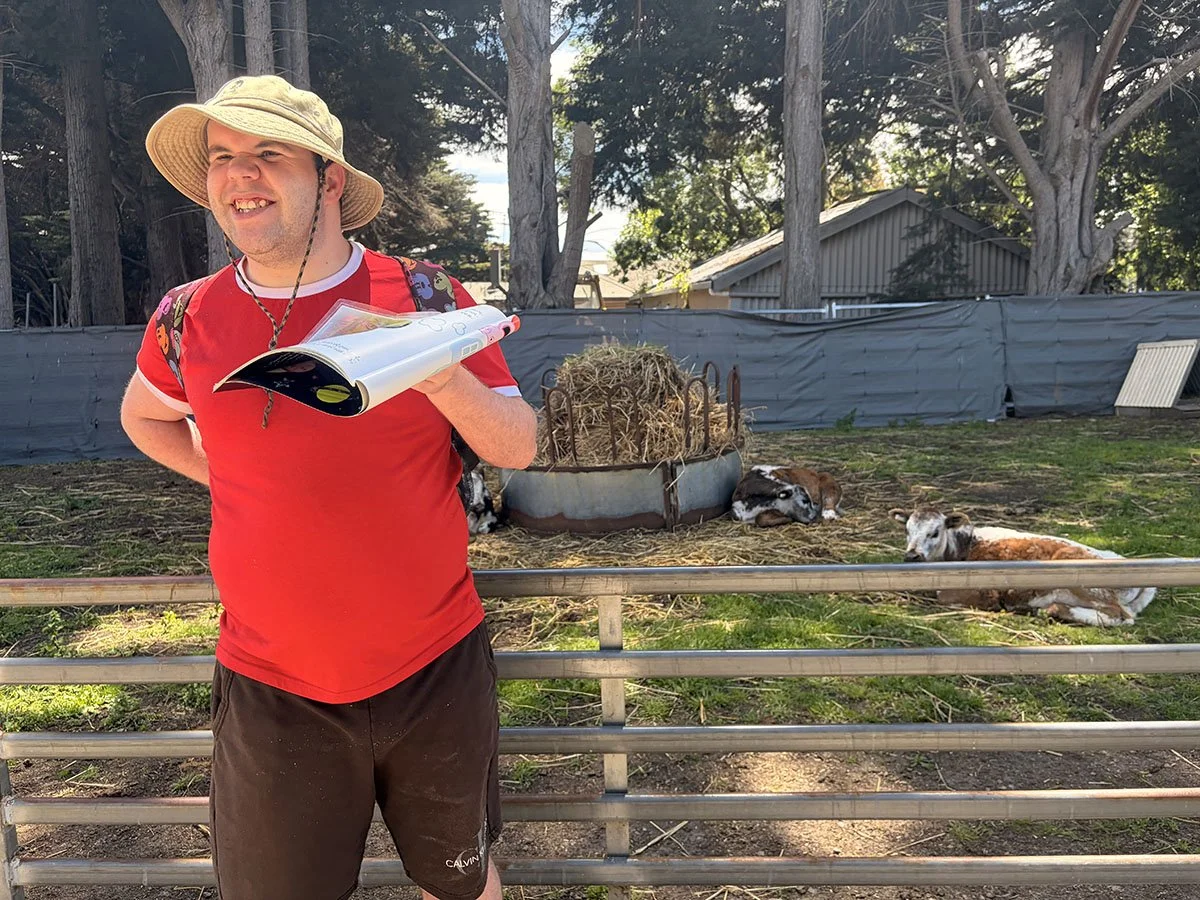 A man wearing a beige sun hat and a red t-shirt with sunglasses hanging from the neck, holding a colorful book or magazine, standing in front of a farm fence with three calves lying on the ground behind him.