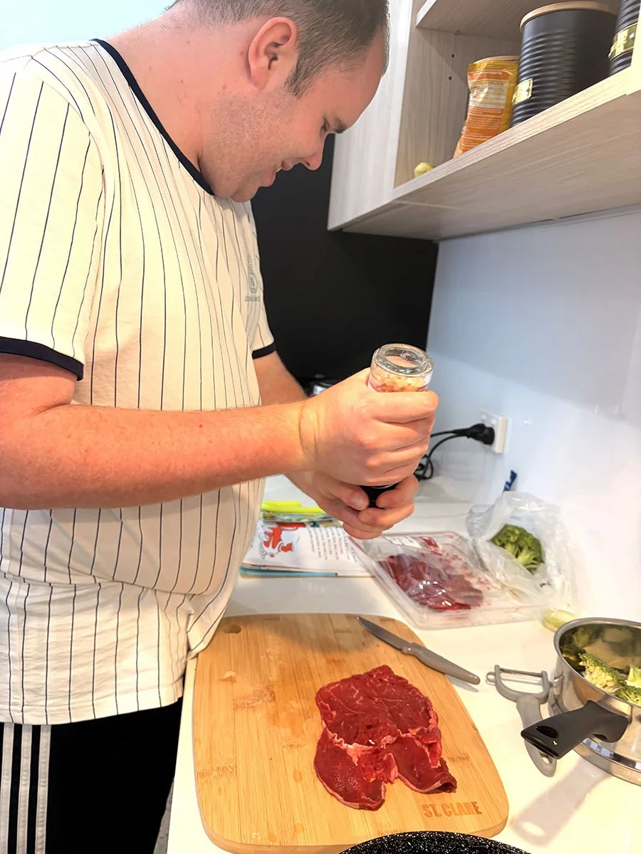 A man preparing to season a raw steak on a wooden cutting board in a kitchen. There are cooking utensils, a knife, and vegetables around him.