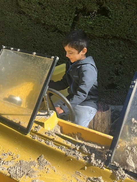 A young boy playing on a yellow sand and water playground structure outdoors, with trees in the background.