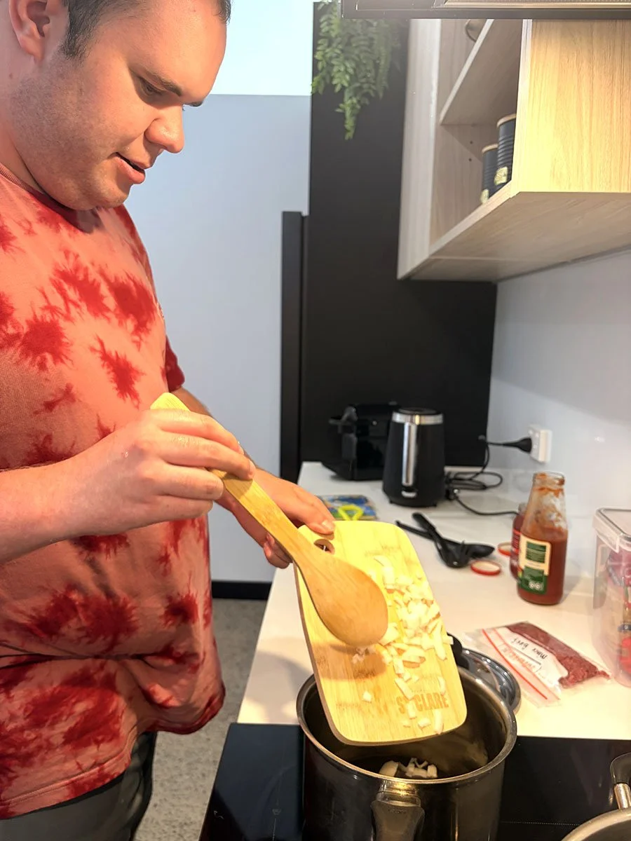 A man in a red tie-dye shirt is stirring ingredients in a pot on a stove while holding a wooden cutting board.