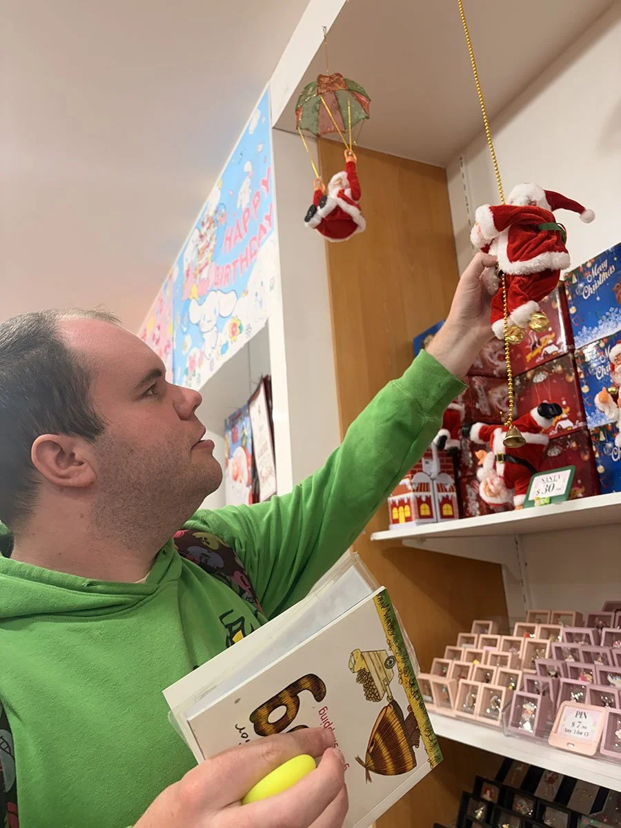 A man in a green hoodie shopping for Christmas decorations at a store, holding a booklet and reaching up to the shelf. The shelves display small Santa figurines, some dressed in red and white outfits, and other Christmas ornaments.