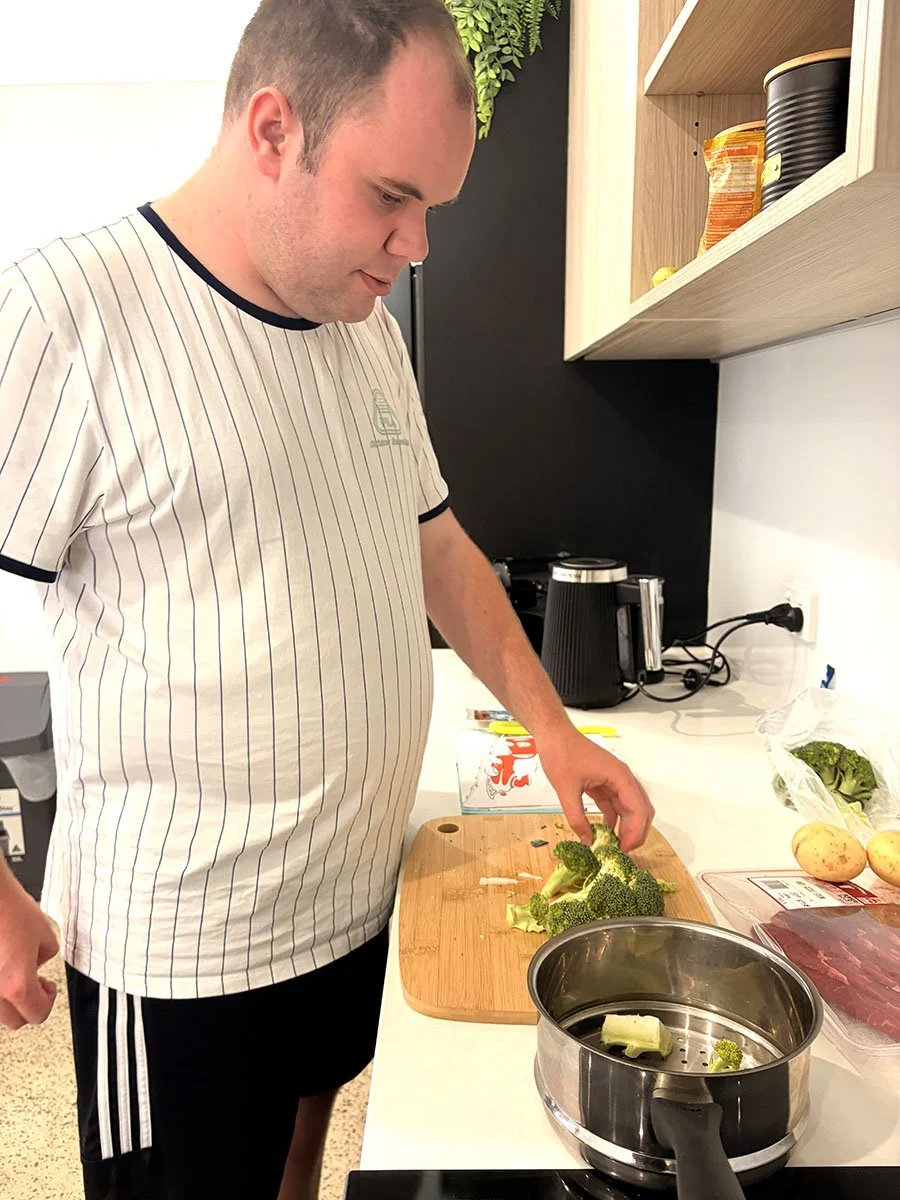 A man is chopping broccoli on a wooden cutting board in a kitchen, with broccoli and potatoes nearby, and a pot on the stove.