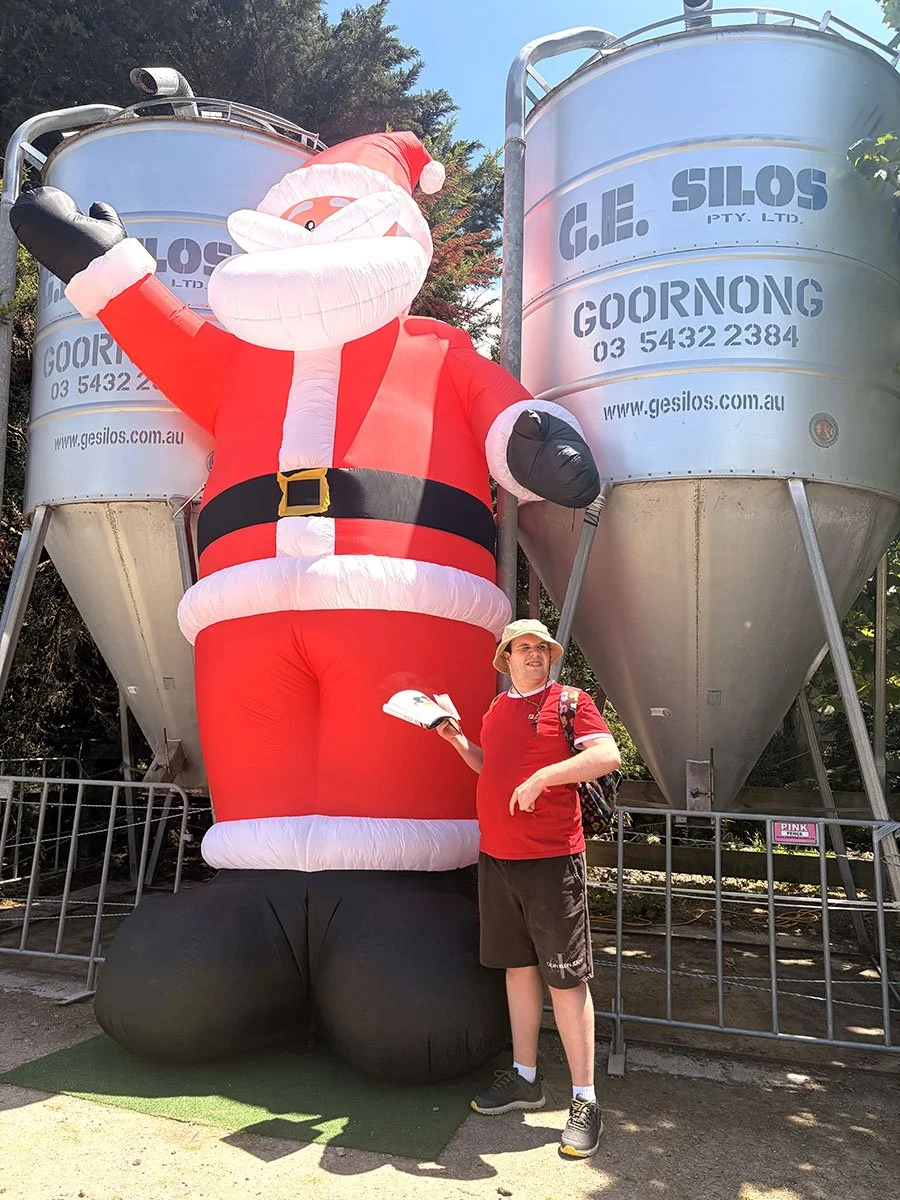 A large inflatable Santa Claus with a waving hand, standing outdoors near metallic silos, with a young man in casual clothes, a red t-shirt, dark shorts, holding a book, and wearing a sun hat.