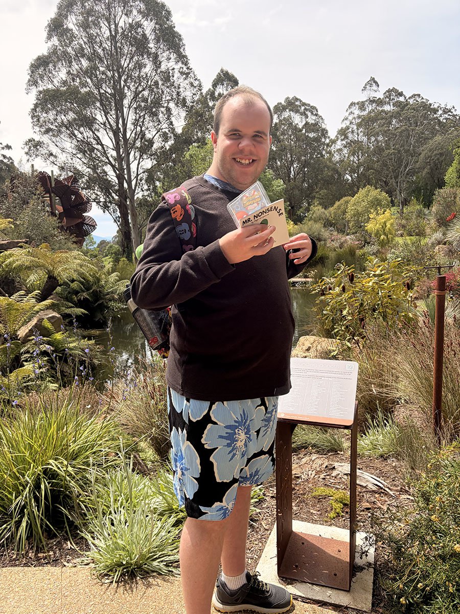 A man standing outdoors near a pond, holding a booklet and a sticker sheet, wearing a black sweatshirt, floral shorts, sneakers, and a backpack with colorful designs. Trees and plants surround him under a cloudy sky.