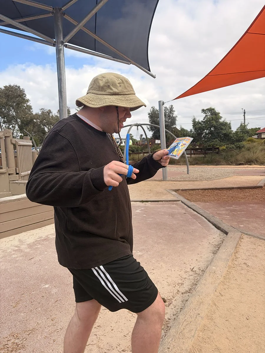 A person wearing a khaki bucket hat, black sweatshirt, and black shorts with white stripes, holding a colorful booklet and a blue jump rope, at a playground under partly cloudy skies.