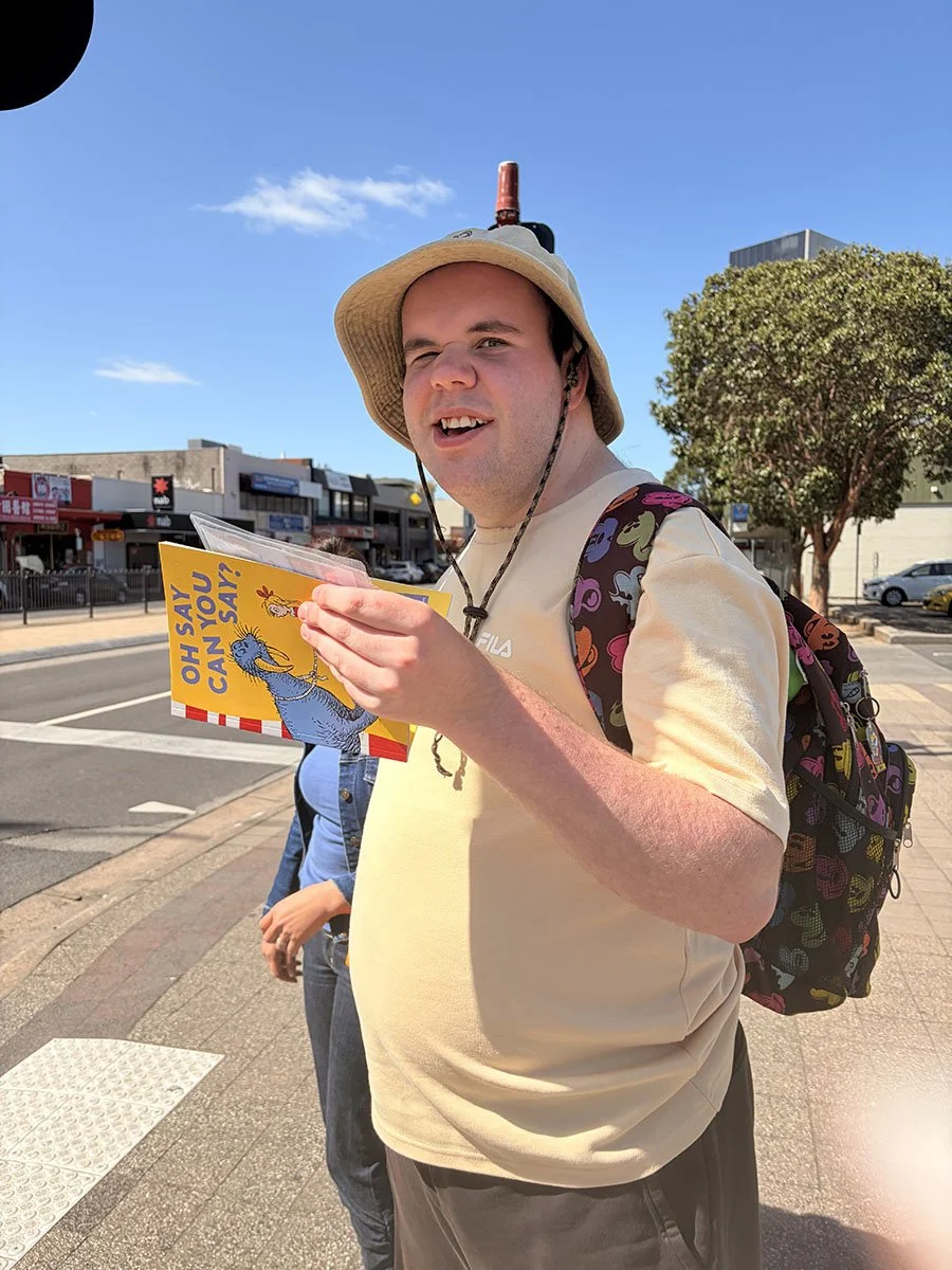 Young man outdoors on sunny day wearing a wide-brim hat, carrying a colorful backpack, holding a pamphlet titled 'Oh Say Can You Say!' with a brightly colored illustration, standing near a sidewalk with shops and trees in the background.