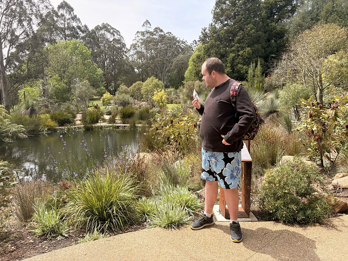 A man standing on a garden path, looking at a small pond surrounded by plants and trees, in a park or botanical garden.
