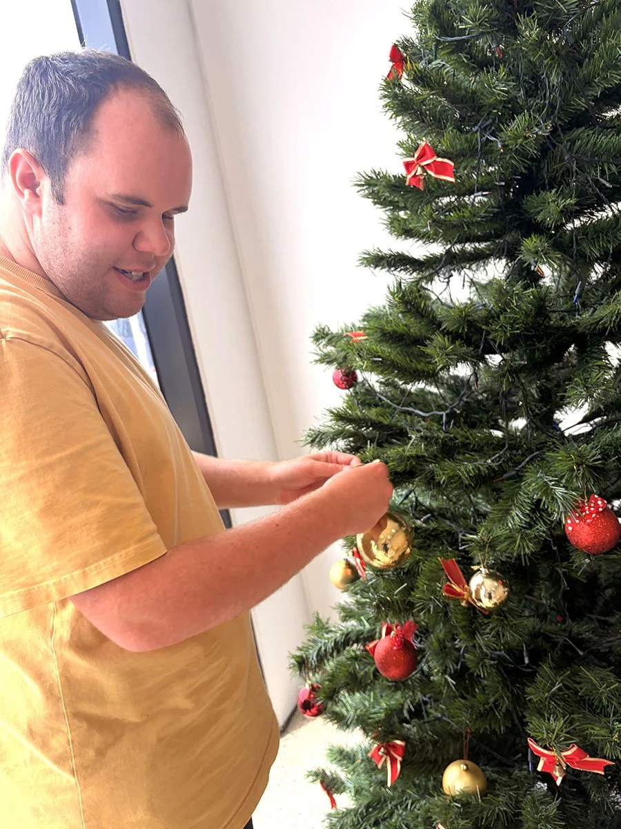A man in a yellow t-shirt decorating a Christmas tree with gold and red ornaments.