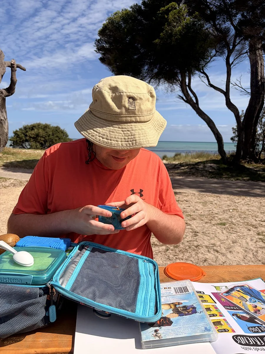 A person wearing a beige bucket hat and red shirt sitting at a picnic table outdoors by the beach, with trees and ocean in the background, looking at a small blue container, with a lunchbox, brochures, and a spoon on the table.