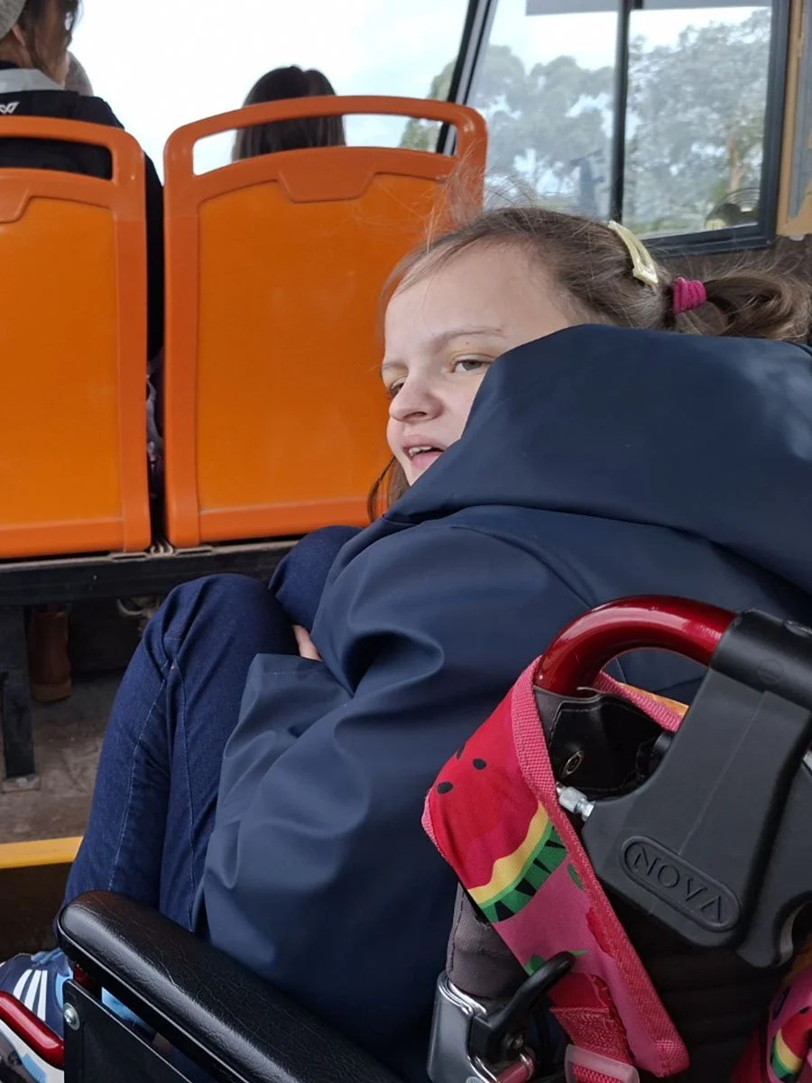 A young girl with red hair sitting on a bus, wearing a navy jacket and jeans, with her arms crossed. The bus has bright orange seats, and windows showing trees outside.