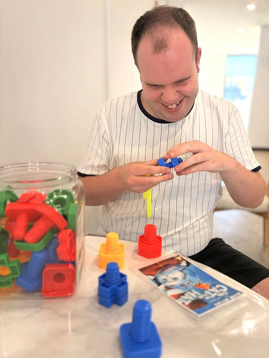 A man playing with colorful Catan game pieces, smiling and assembling a blue game piece. A jar filled with red, green, yellow, and blue game pieces is on the table, along with a Catan rulebook.
