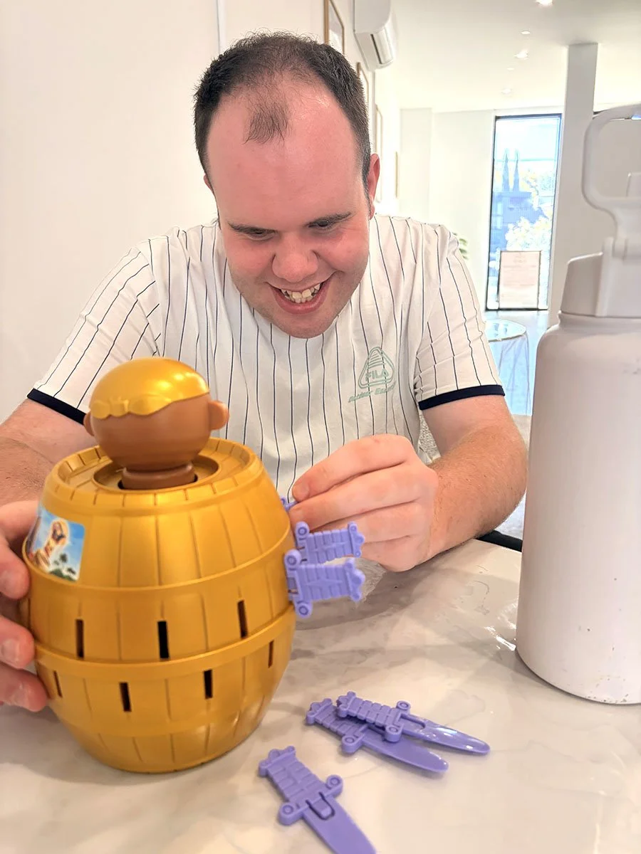 A man smiling and playing with a plastic toy that resembles a golden egg with a small figure of a child with blonde hair on top. The toy is yellow with black lines, and purple plastic pieces are on the table nearby. The man is wearing a white shirt w