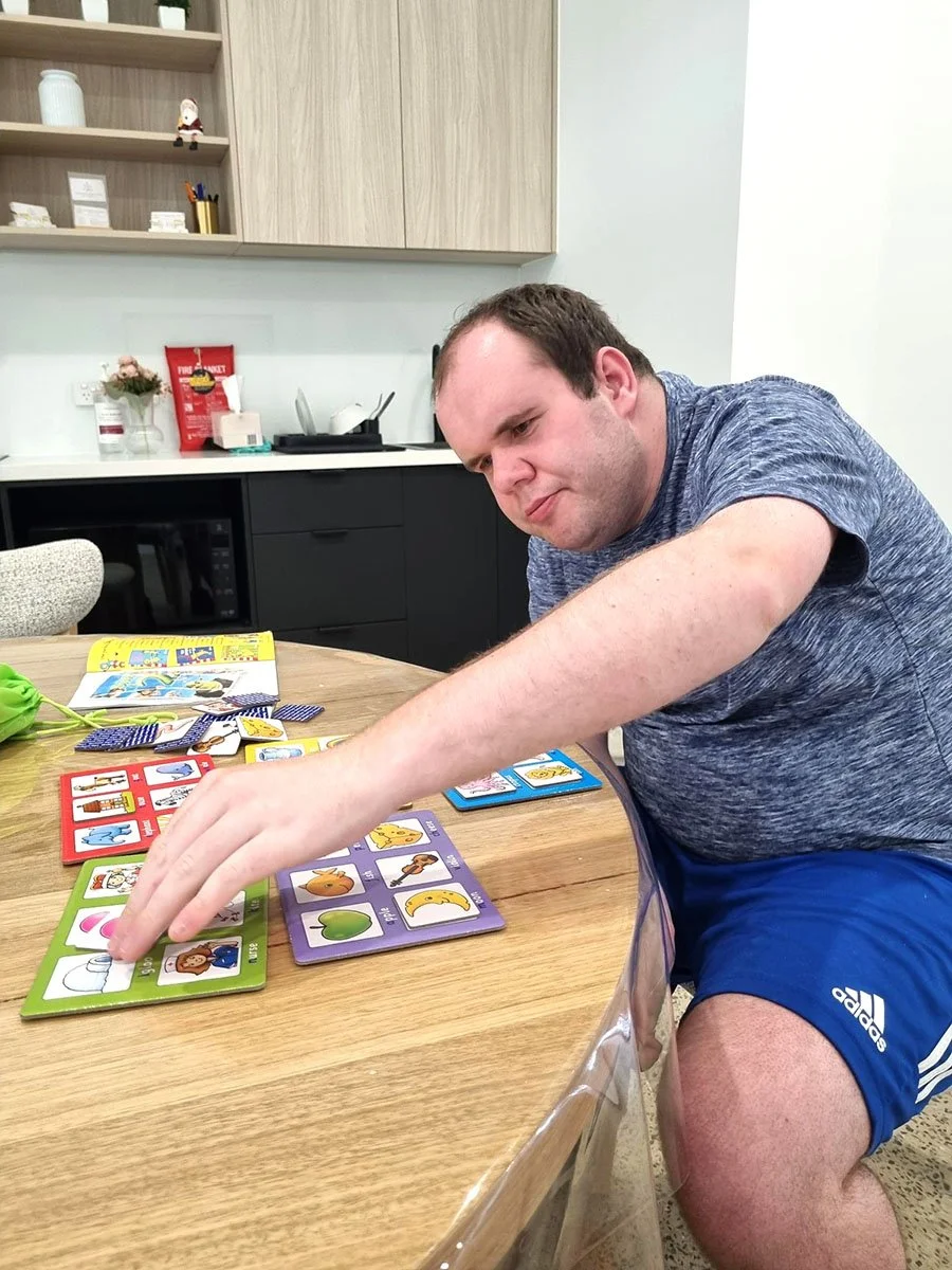 A man sitting at a wooden table playing a card game with colorful cards featuring animals and objects, in a kitchen or break room with shelves and kitchen appliances in the background.