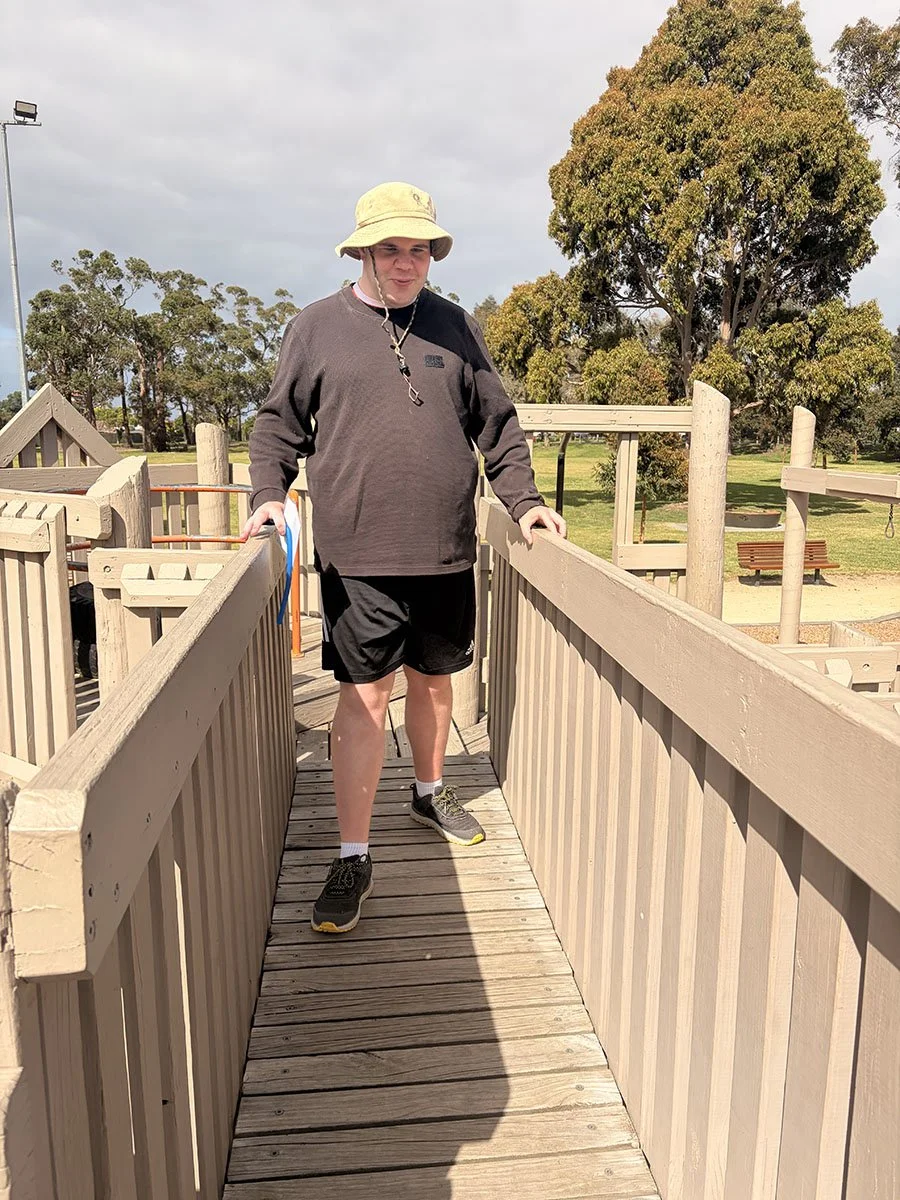 A man wearing a yellow sun hat, black sweatshirt, black shorts, and running shoes stands on a wooden bridge at a park, with trees and open grassy areas in the background.