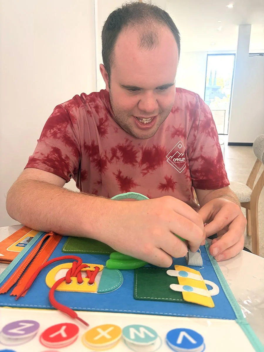 A man with short dark hair and a beard, wearing a red tie-dye t-shirt, is sitting at a table and playing with a colorful felt activity board.