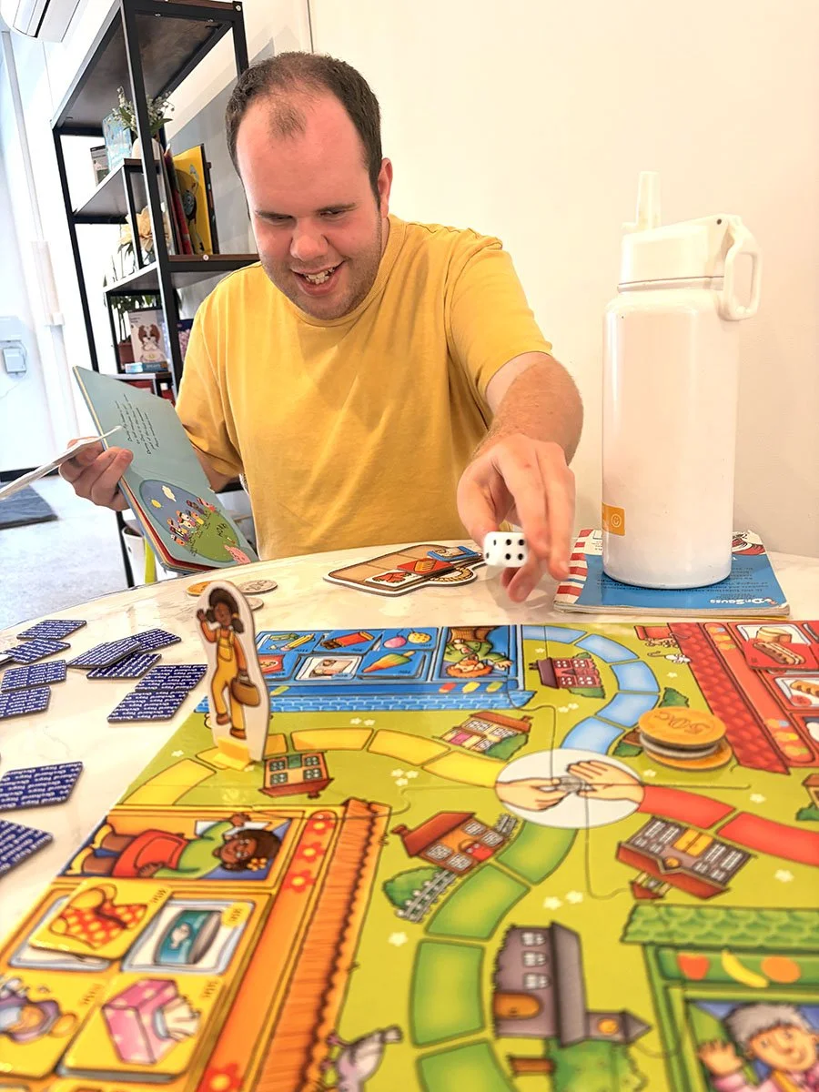 A man in a yellow shirt playing a colorful board game at a table, holding a die and a card, with a water jug in the background.