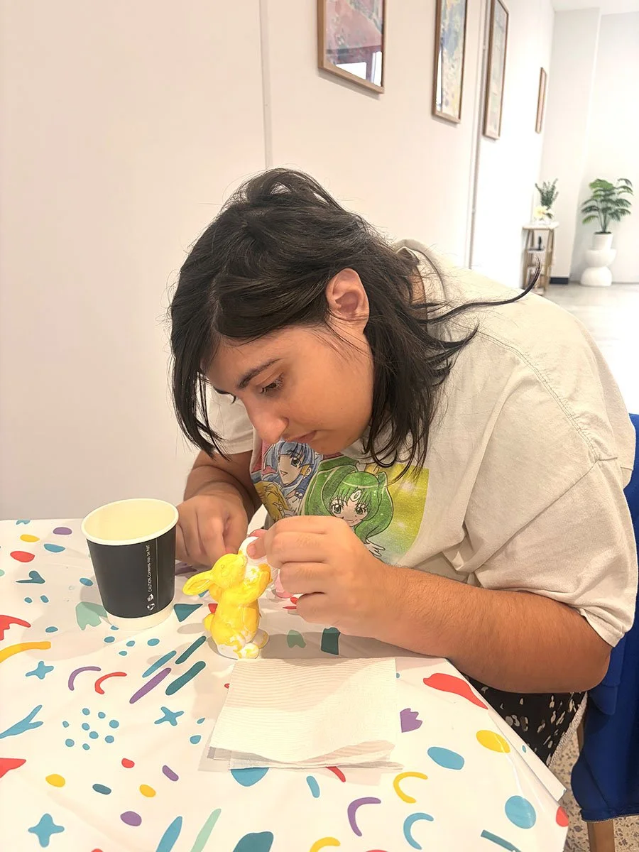 A young woman with dark hair sits at a table decorating a yellow bunny-themed cake. There is a black paper cup and a napkin on a colorful, festive tablecloth with abstract shapes. The background shows a white wall with framed pictures and a potted pl