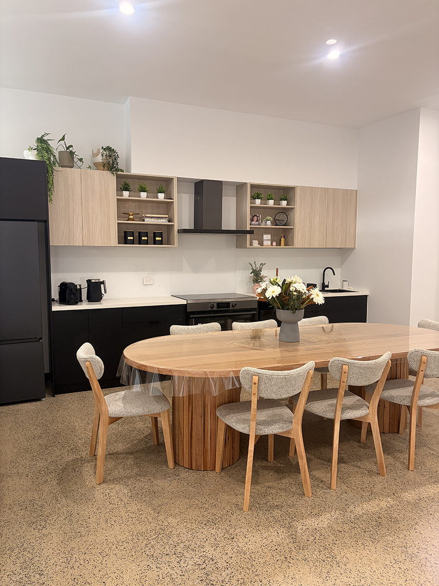 Modern kitchen with a large wooden dining table, six upholstered chairs, black and light wood cabinetry, and decorative plants.