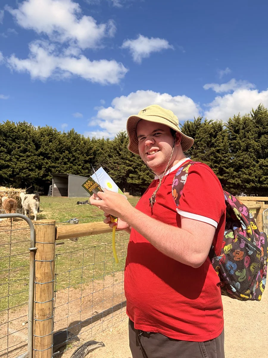 A young man wearing a beige bucket hat, red T-shirt, and a colorful backpack stands outdoors near a fenced enclosure with animals, holding a pamphlet and a yellow balloon on a string. The sky is blue with some clouds, and trees are in the background.