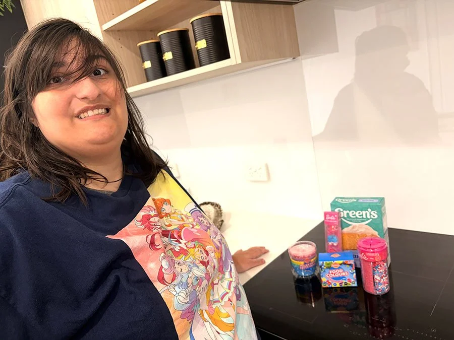 A woman with dark hair and glasses is smiling at the camera in a kitchen. On the black countertop, there are colorful candies and snacks, including a box of Green's cake mix, a pack of Skittles, and a jar of sprinkles.