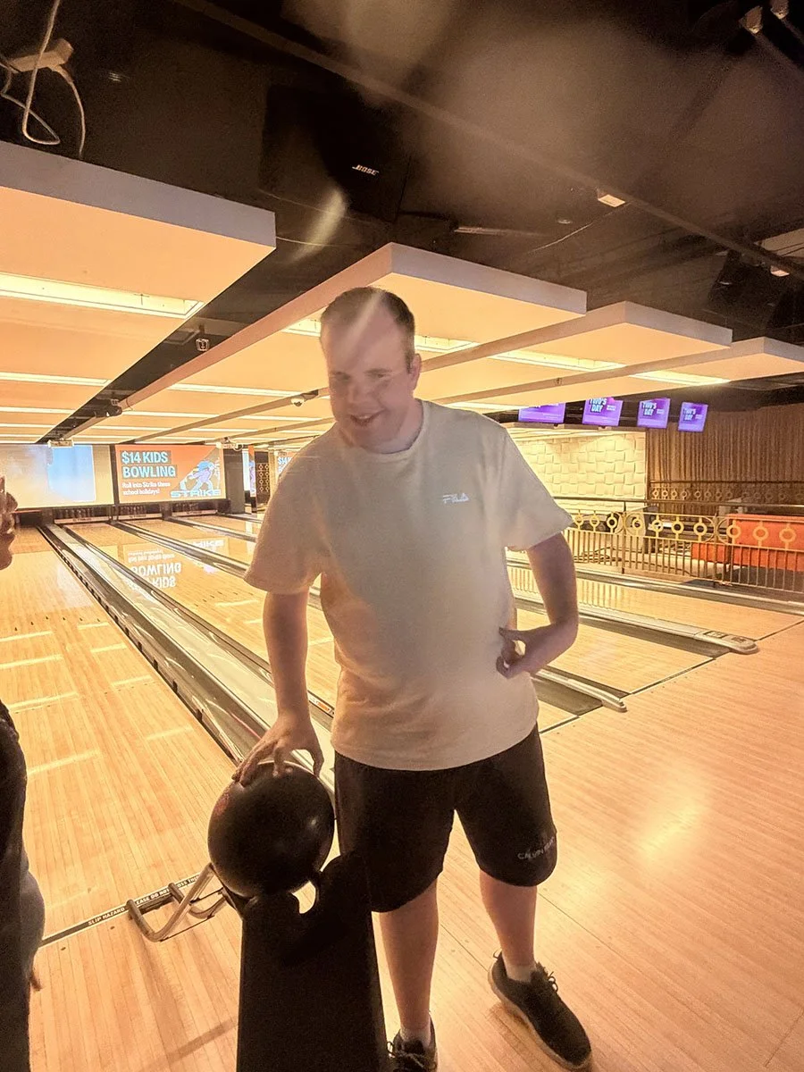 A young man at a bowling alley holding a bowling ball in one hand and smiling.