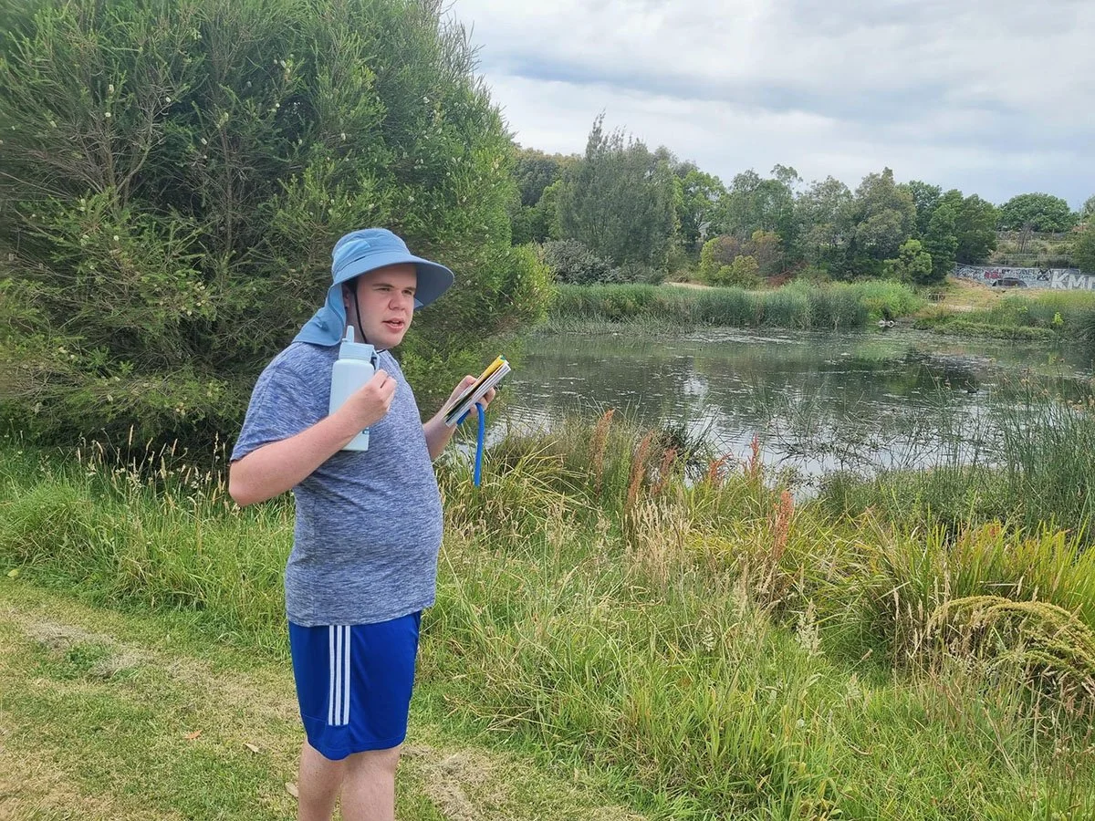 A boy standing outdoors near a pond, holding a water bottle and a small notebook, wearing a blue sun hat and casual clothes.
