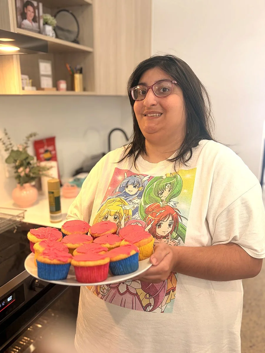A woman with glasses holding a plate of colorful cupcakes decorated with pink icing in a kitchen.