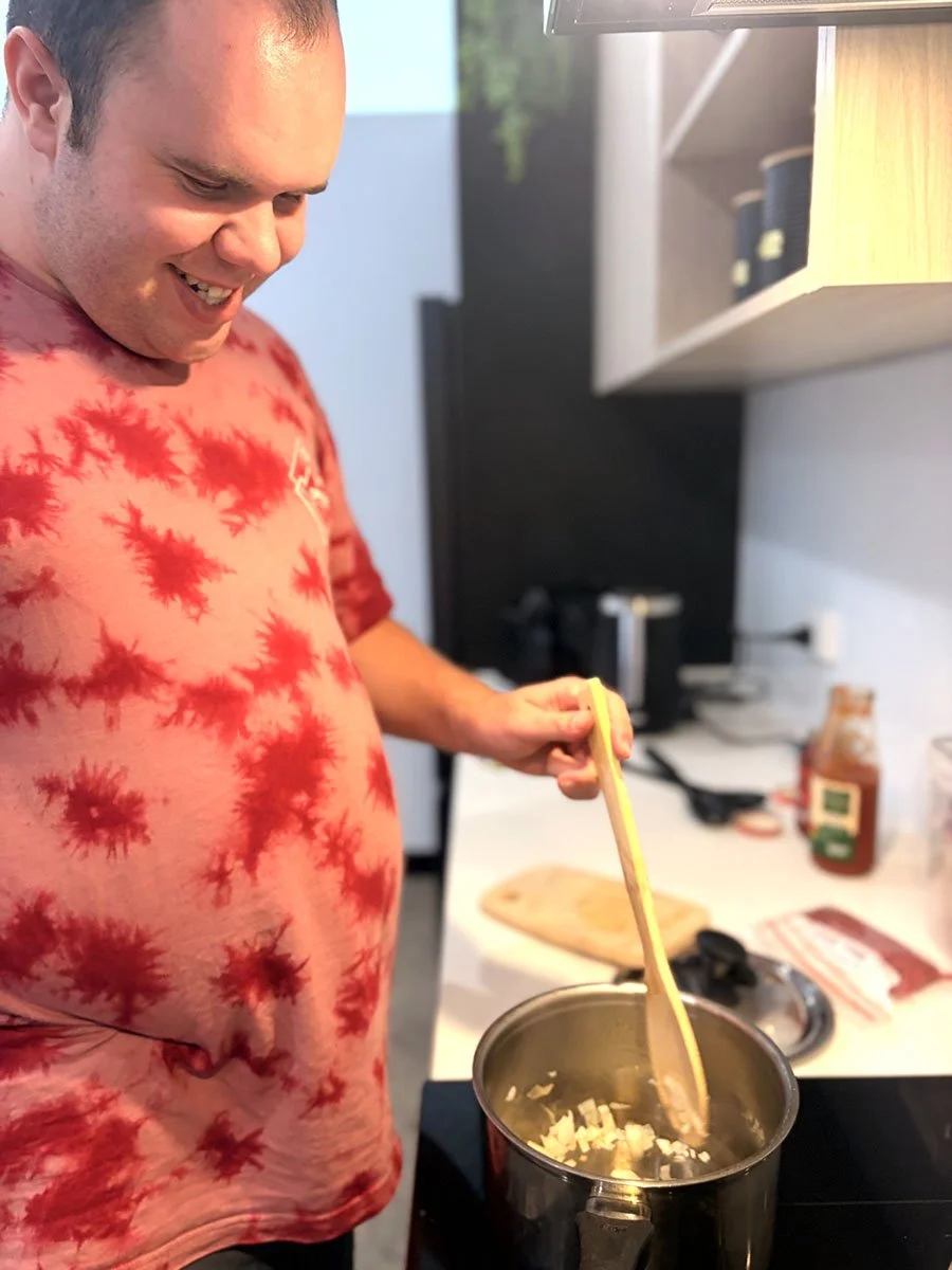 A man wearing a red tie-dye t-shirt is stirring a pot of chopped onions on a stove in a kitchen.