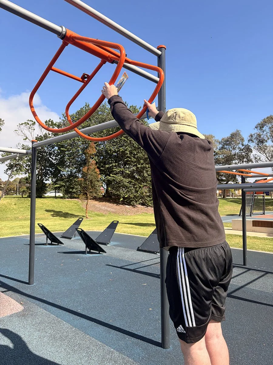 A person wearing a tan bucket hat, dark jacket, and black Adidas shorts, reaching up to adjust or hold onto a hanging orange climbing structure at a playground. The background features a grassy park with trees, blue sky, and blue rubberized playgroun