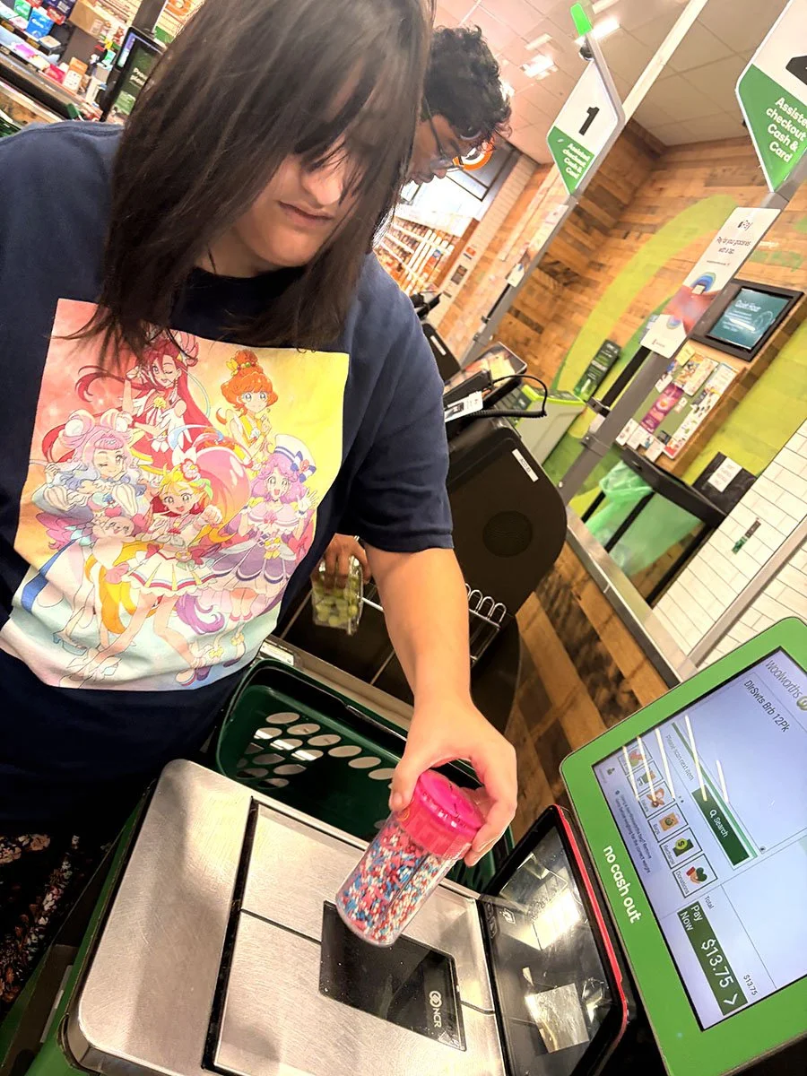 A woman shopping at a grocery store checkout, placing a container of sprinkles on the scanner. She is wearing a dark sweatshirt with colorful anime-style characters on it. In the background, a man is visible, and store signage and checkout counters a