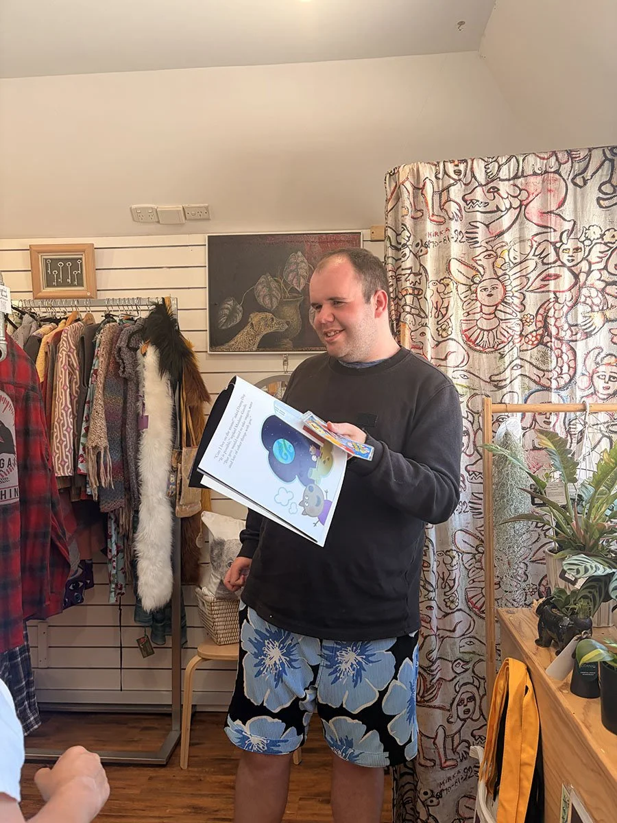 A man standing inside a clothing store holding a colorful book, smiling and looking at someone off-camera. He is wearing a black sweatshirt and blue floral shorts. Behind him are folded plants, a curtain with colorful doodles, and clothing racks with