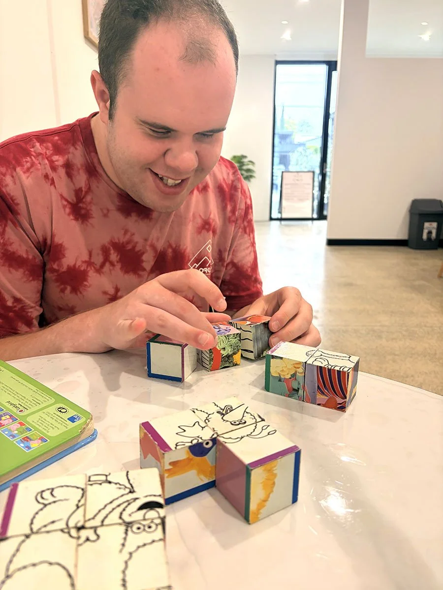 A man in a red tie-dye shirt playing with colorful, illustrated paper cubes on a white table inside a well-lit room.