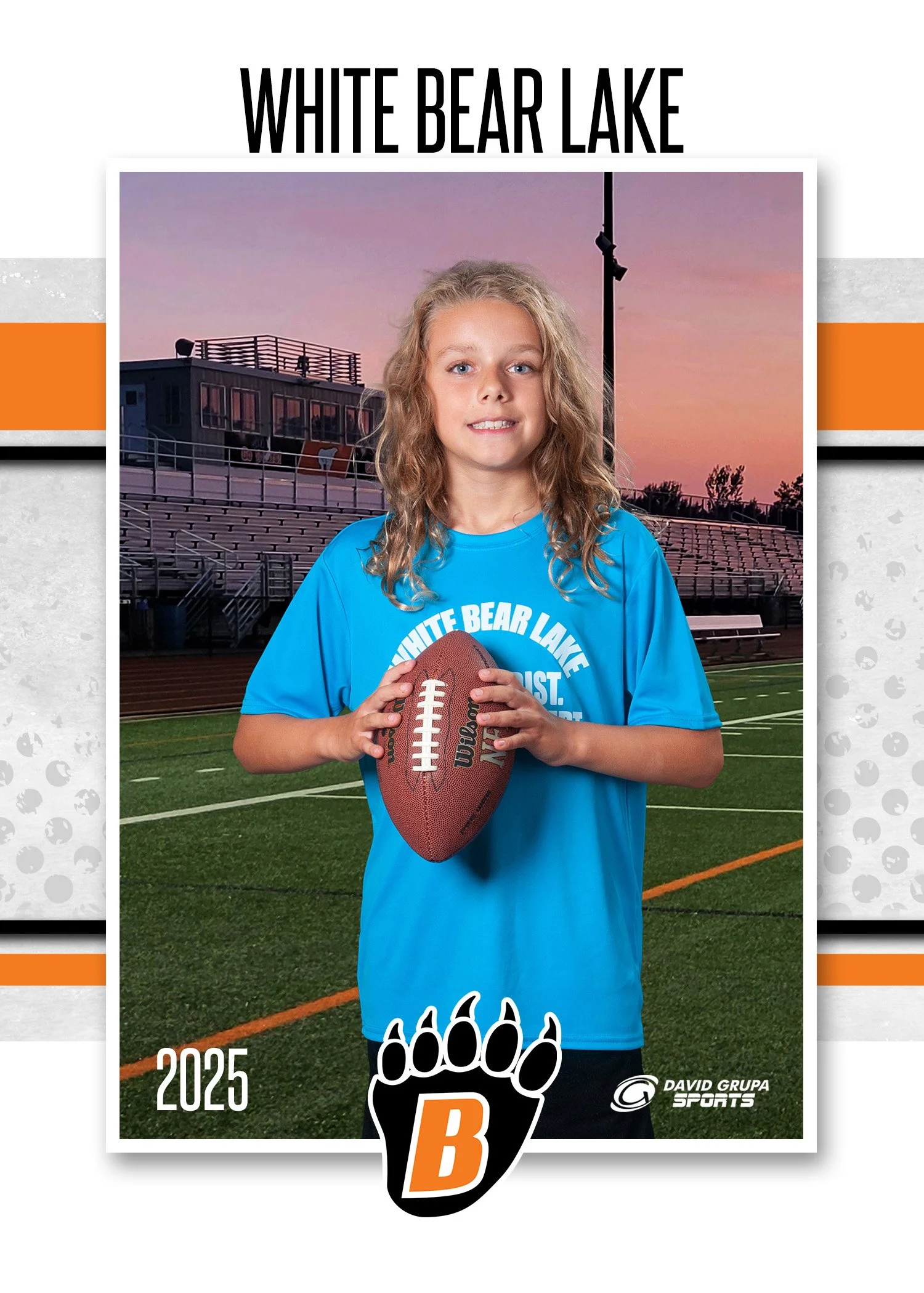 A young girl with curly blonde hair holding a football on a football field during sunset. She is wearing a blue shirt with the words 'White Bear Lake' and a logo with a paw print and a letter B. The background shows bleachers and a press box at the s