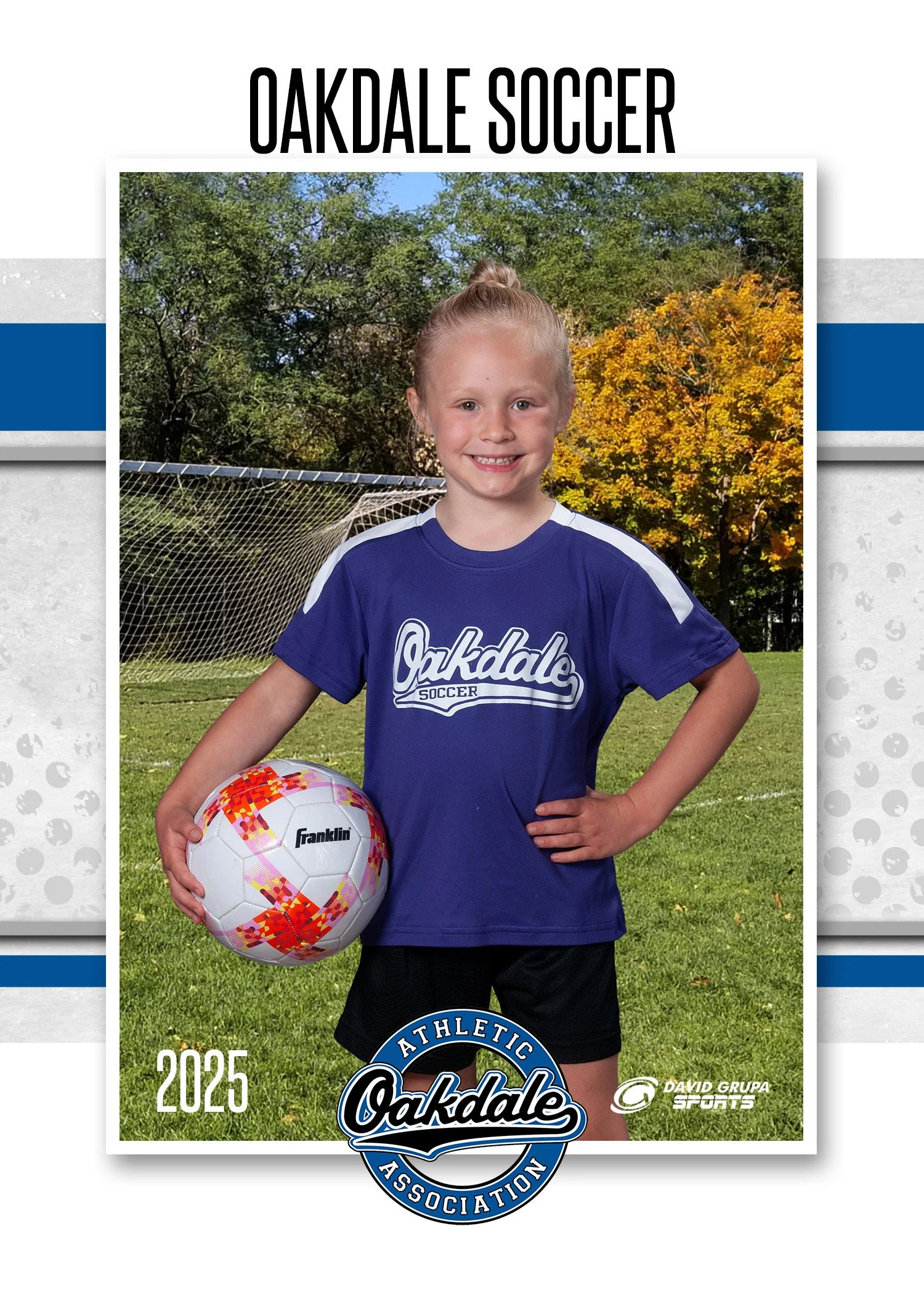 Young girl in blue Oakdale Soccer jersey holding a white soccer ball with pink and orange patterns on a soccer field with trees in the background, smiling at the camera.