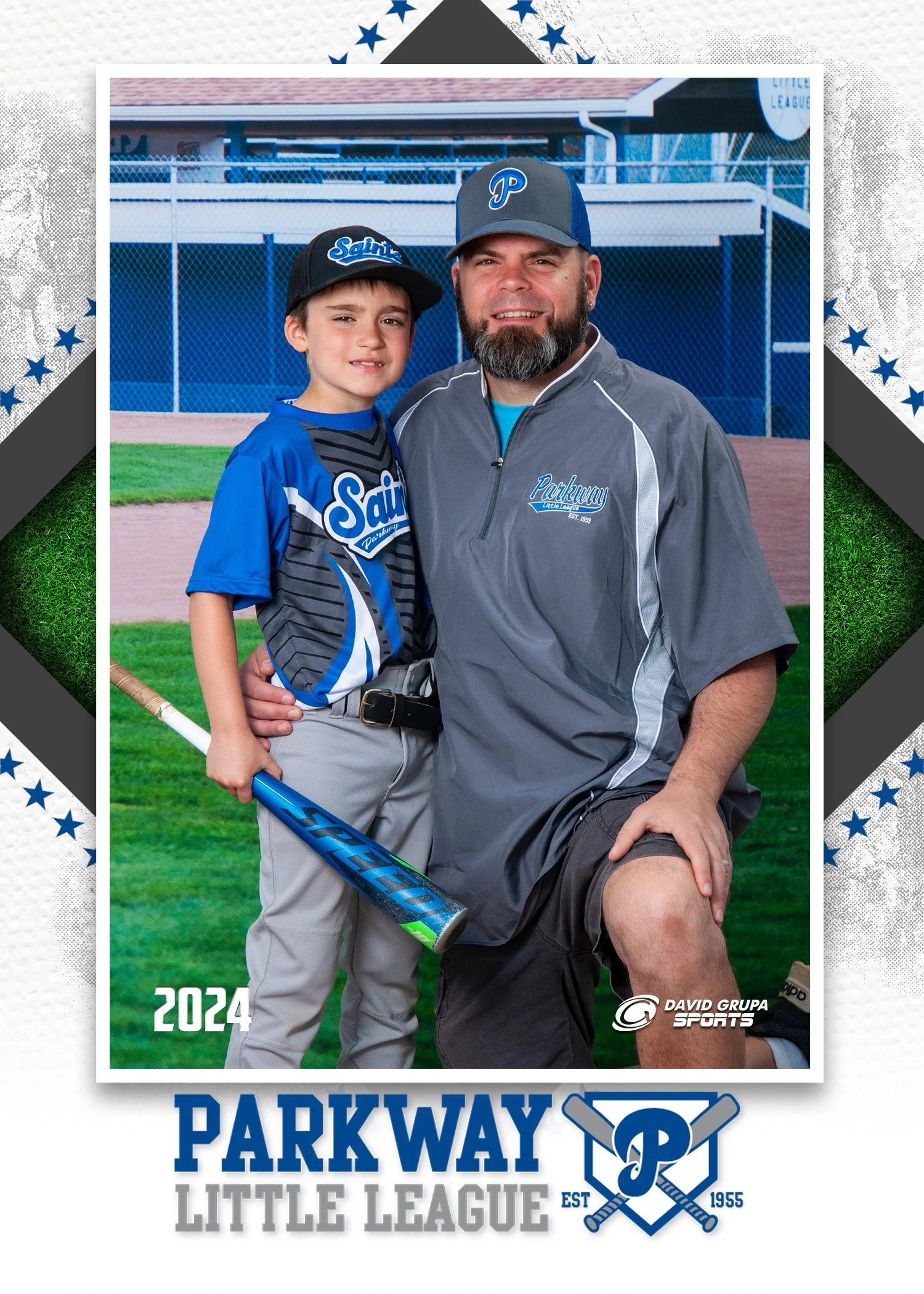 Photograph of a young boy and an adult man, likely father and son, together on a baseball field. The boy is dressed in a blue and gray baseball uniform, holding a bat, and wearing a cap that says 'Saints.' The man is dressed in a gray and blue jacket