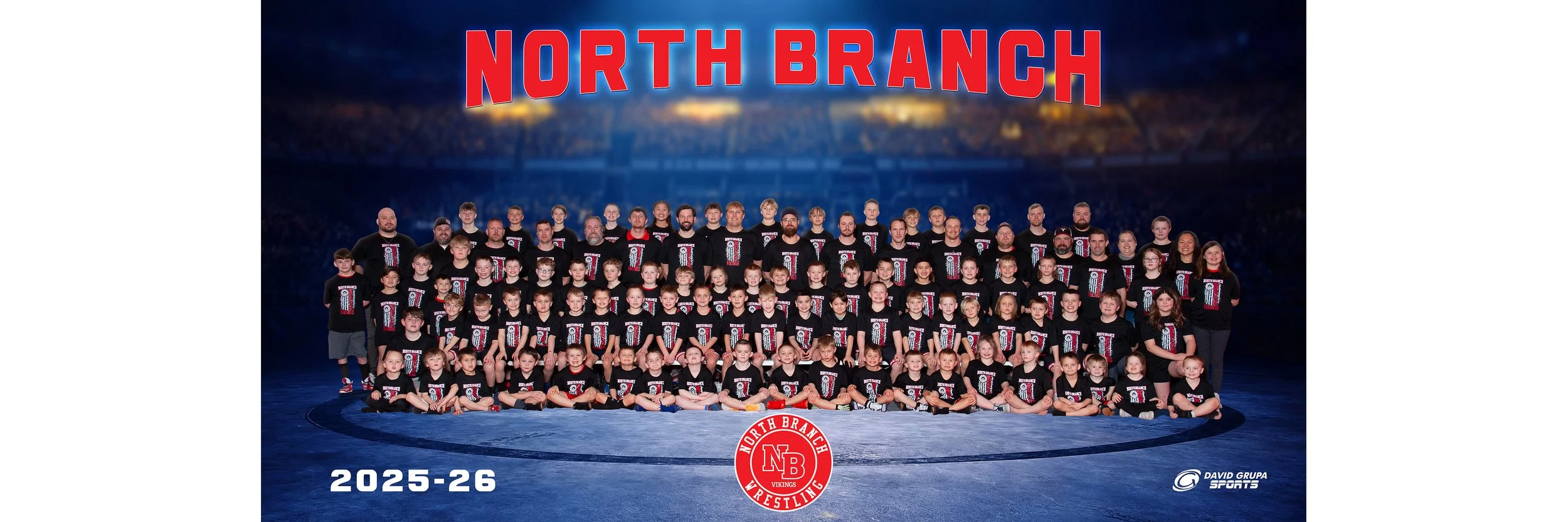 Group photo of North Branch wrestling team members, including children and adults, standing on a wrestling mat in a sports arena. The team is wearing black shirts with the team logo. The background features a large sticker with the team name, and the photo indicates the 2025-26 season.